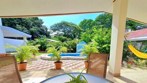 A covered patio with a glass table and wicker chairs, surrounded by potted plants and a swimming pool.