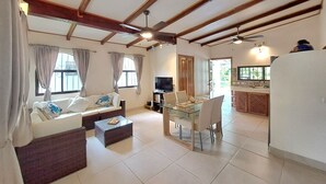 A spacious living room with a sofa, coffee table, and dining area. A television is on a stand, and there is a kitchen area visible in the background.