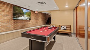 A pool table with a red felt surface, a television mounted on a brick wall, and a sofa with a cow-patterned pillow.