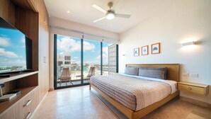 A modern bedroom with a large bed, a ceiling fan, a TV, and a balcony with a view of the city. - 1 Bedroom Pool & Gym - Piedra Azul 605 (Playa del Carmen)