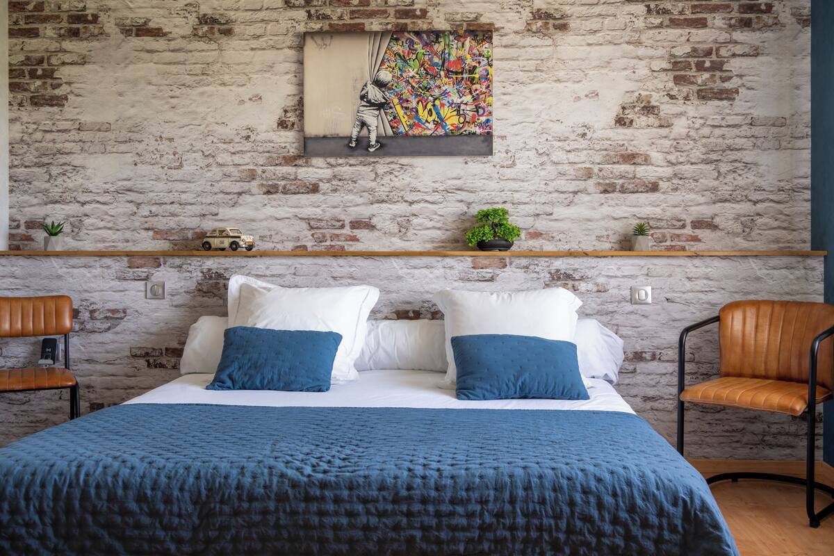 A bedroom with a brick wall, a bed with blue bedding, two chairs, and a shelf with plants and a toy car.