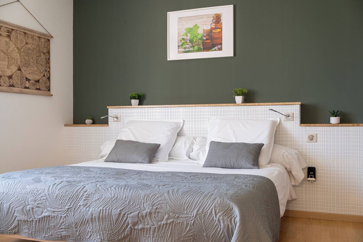 A neatly made bed with a grey patterned bedspread, white pillows, and a wooden headboard with small potted plants.