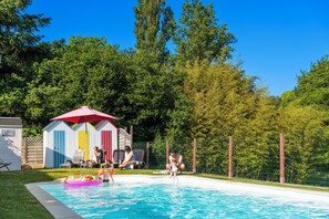Une piscine avec un parasol rouge, une cour clôturée et, au fond, des abris de jardin aux couleurs vives.