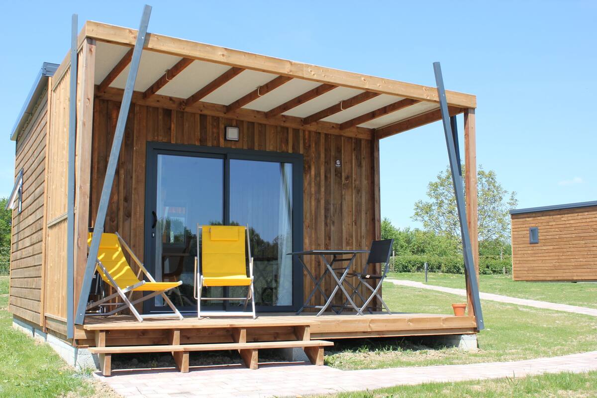 A wooden cabin with a porch, two yellow chairs, a table, and a sliding glass door.