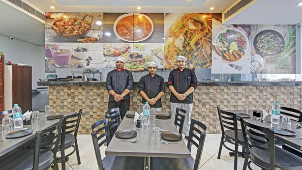 Interior de restaurante con tres cocineros frente a una pared de imágenes de comida, mesas dispuestas con platos y vasos, y un salpicadero de azulejos.