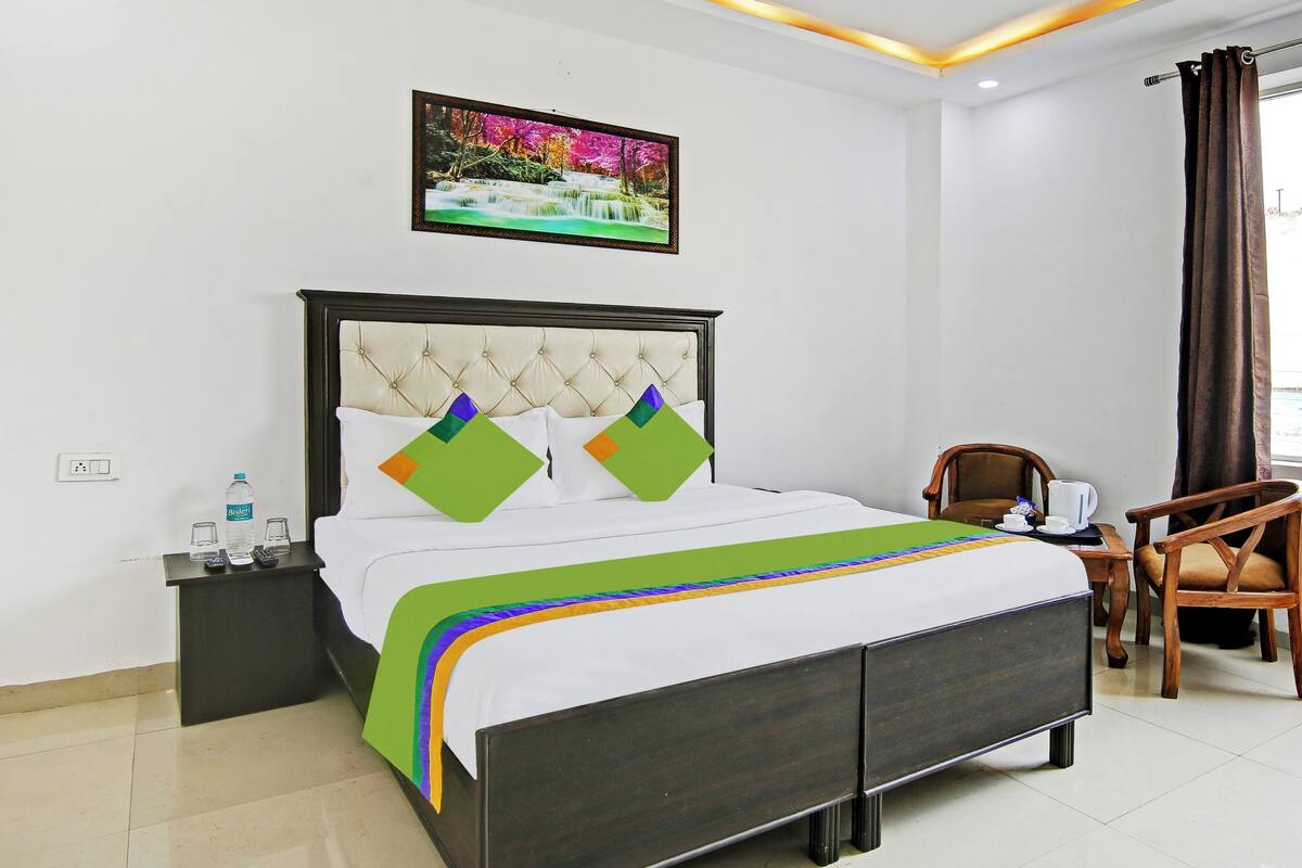 Habitación de hotel con cama, mesita de noche, una silla, ventana con cortinas y un cuadro en la pared.