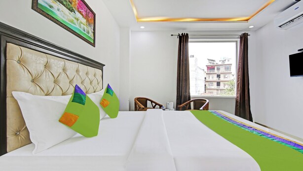 Een hotelkamer met een gestoffeerd hoofdeinde, een bed met witte beddengoed en kleurrijke kussens, een stoel, een raam met gordijnen en een flatscreen-televisie.