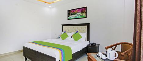 A hotel room with a bed, a bedside table with a phone, a chair, and a table with a kettle and cups.