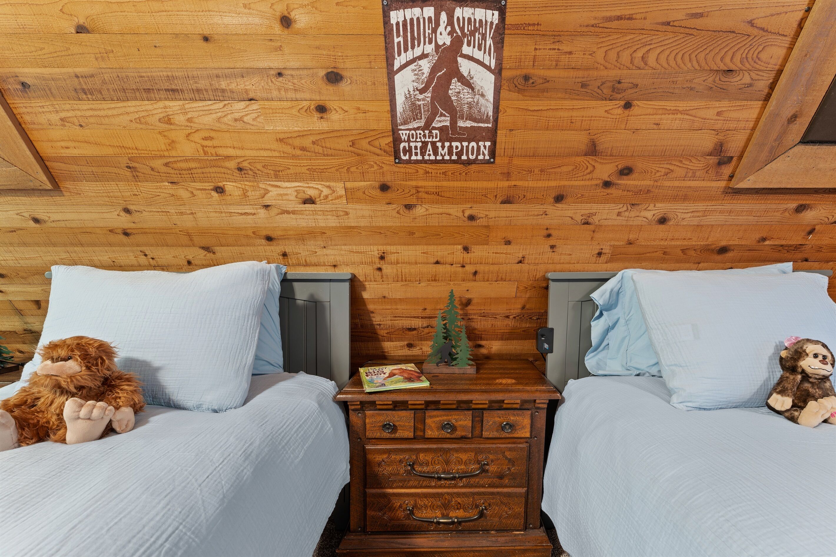 Techo de madera con un letrero que dice 'Campeón Mundial de Escondite', dos camas con ropa de cama azul claro, una mesita de noche de madera con un libro y una pequeña decoración de árbol, y un osito de peluche sobre una de las camas.