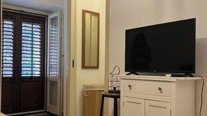 Una habitación con una televisión sobre un mueble blanco, una puerta con persianas y un espejo en la pared.