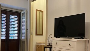 Un dormitorio con cama, un televisor sobre un mueble blanco, un espejo y una puerta que da a otra habitación.