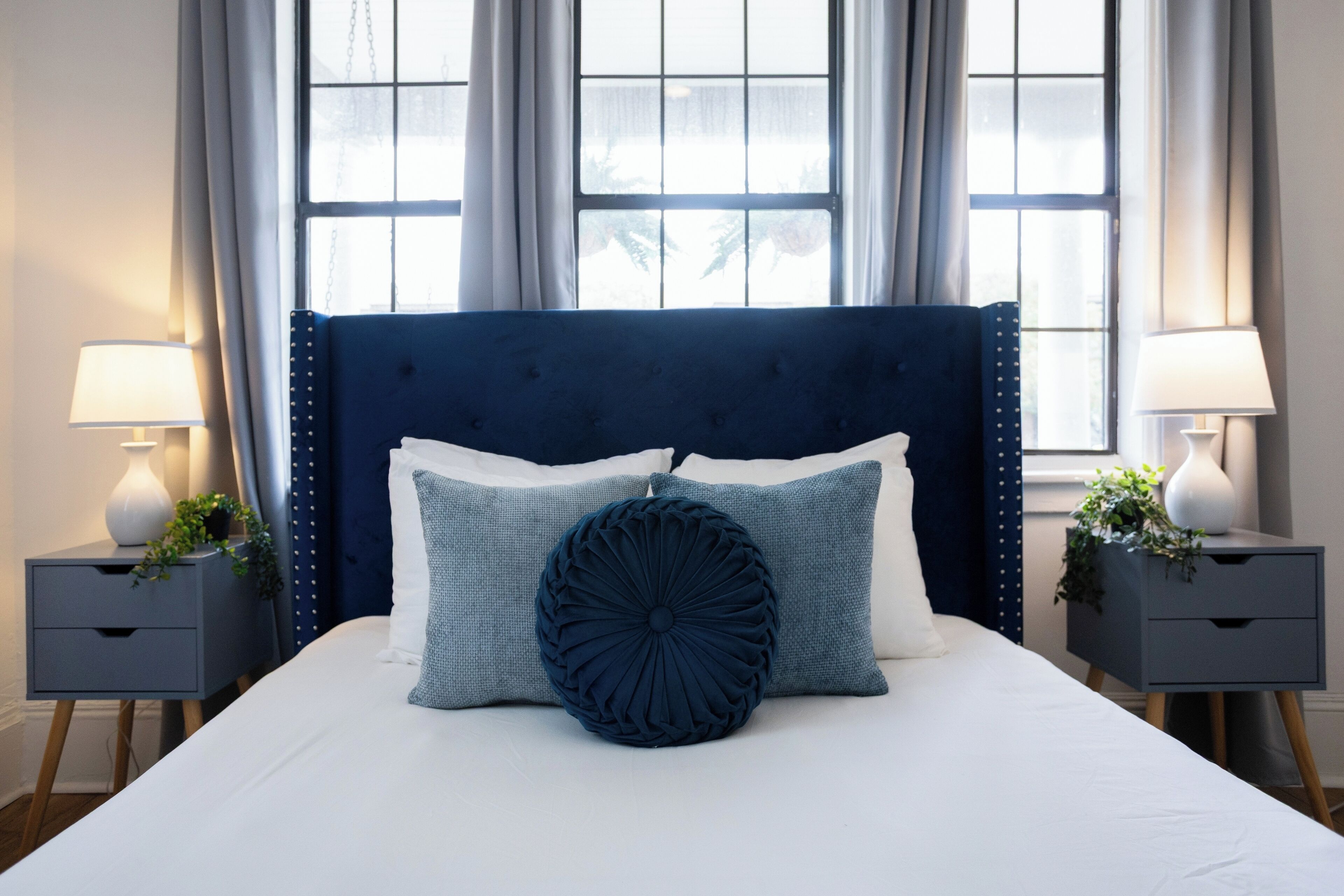 Uma cama bem arrumada com cabeceira azul escura, duas mesas de cabeceira com luminárias e vista para o exterior por grandes janelas.
