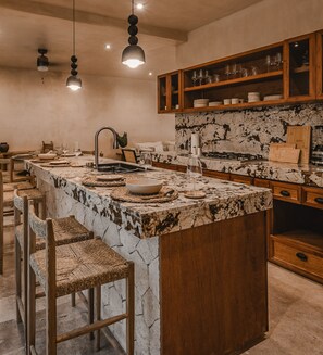 Une cuisine dotée d’un îlot central en marbre, de tabourets de bar en bois et d’une étagère supérieure en bois où sont rangés des verres.