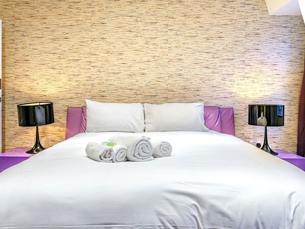Un lit bien fait, avec du linge de lit blanc et des serviettes pliées, deux lampes de chevet et un mur à texture.