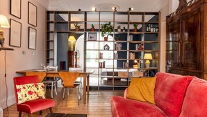 Un salon chaleureux avec un canapé rouge, une table basse en bois et un coin repas comprenant une table et des chaises en bois. On y trouve une bibliothèque remplie de livres et d’objets décoratifs, ainsi qu’une grande armoire dans un coin.