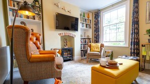 A cozy living room with a leather armchair, a yellow ottoman, a fireplace, a bookshelf, and a television. - Pass the Keys | Charming 2 Bed Townhouse Short Walk To The River (Shrewsbury)