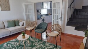 A living room with a sofa, two wicker chairs, a small table with a plant, and a green patterned rug. A kitchen with white cabinets and a washing machine is visible in the background.