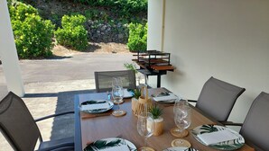 A dining table set with plates, cutlery, and glassware, surrounded by chairs, with a view of greenery outside.