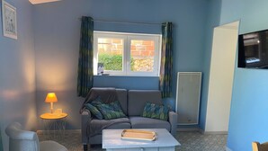 A cozy room with a grey sofa, a small table, and a chair. There's a window with curtains, a radiator, and a TV mounted on the wall. - Gîte Les Lauzes "A precious interlude in the heart of the Périgord Noir". (Saint-Avit-de-Vialard)