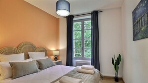 A neatly made bed with pillows and towels, a bedside lamp, a window with curtains, and a potted plant. - Nuit Étoilée - Cocoon in the Heart of Arles - A/C (Arles)