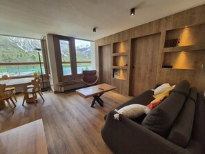 Uma sala de estar moderna com piso de madeira, um sofá, uma área de jantar com cadeiras e vista para montanhas através de grandes janelas.