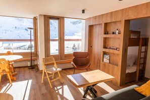 Quarto moderno com piso de madeira, uma mesa redonda, uma cadeira marrom, um sofá e uma estante de livros. Grandes janelas revelam uma vista de montanhas nevadas.