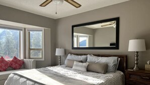 Ein Schlafzimmer mit einem großen Bett, zwei Nachttischlampen, einem Deckenventilator, einem großen Spiegel und einem Fenster mit Blick auf Bäume.