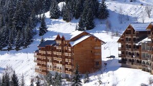 A multi-story wooden lodge with balconies, surrounded by snow-covered trees and a mountainous backdrop.
