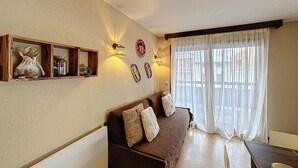 Un salon chaleureux avec un canapé marron, une étagère en bois ornée d’objets décoratifs, une horloge murale et une grande fenêtre avec des rideaux légers.