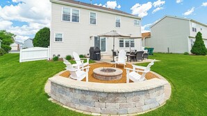 Ein Garten mit einem Steinkamin, weißen Adirondack-Stühlen und einem Patio-Tisch mit Sonnenschirmen.
