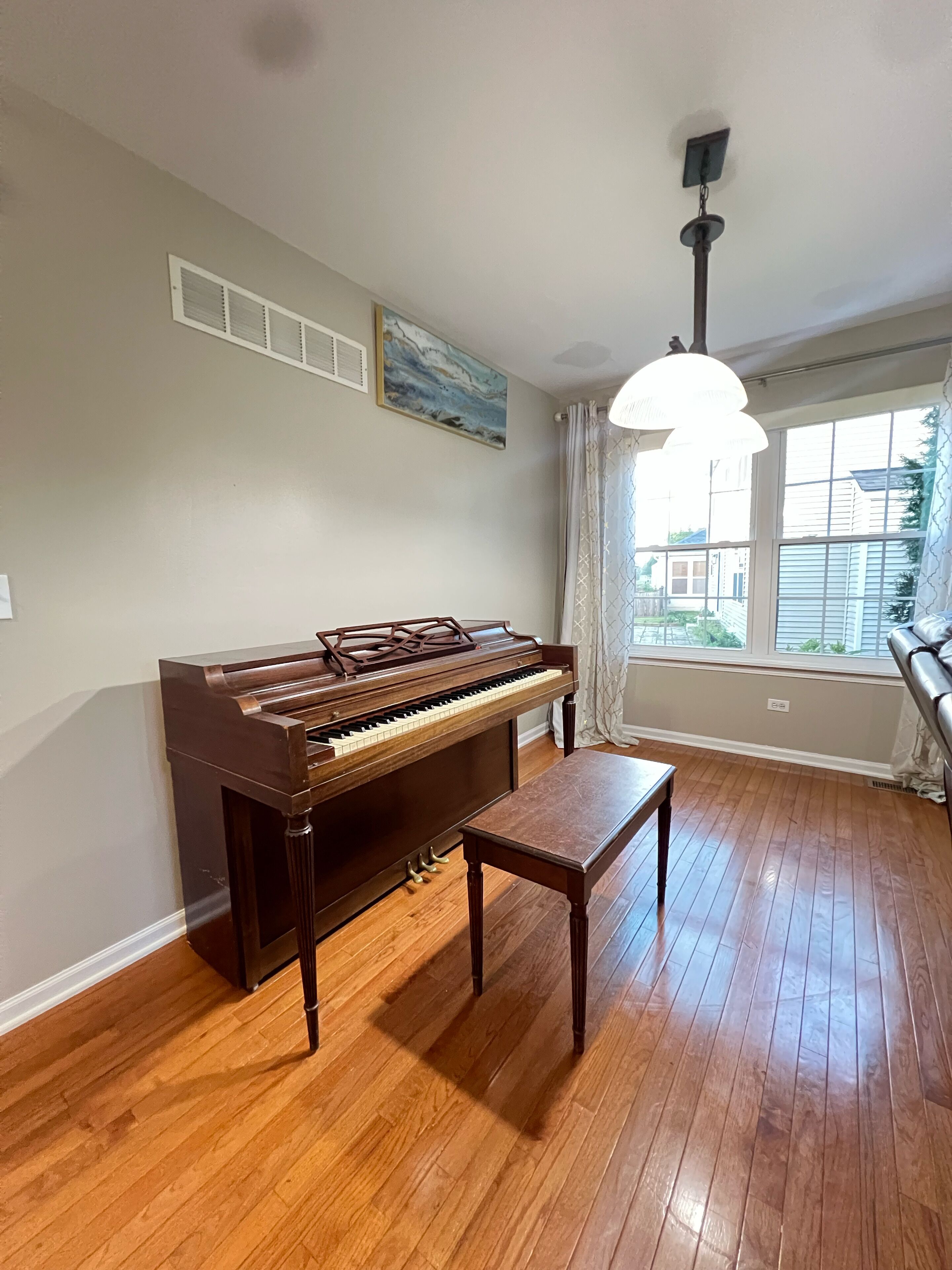 Een kamer met een houten piano, een tafel, een schilderij en een raam met gordijnen.