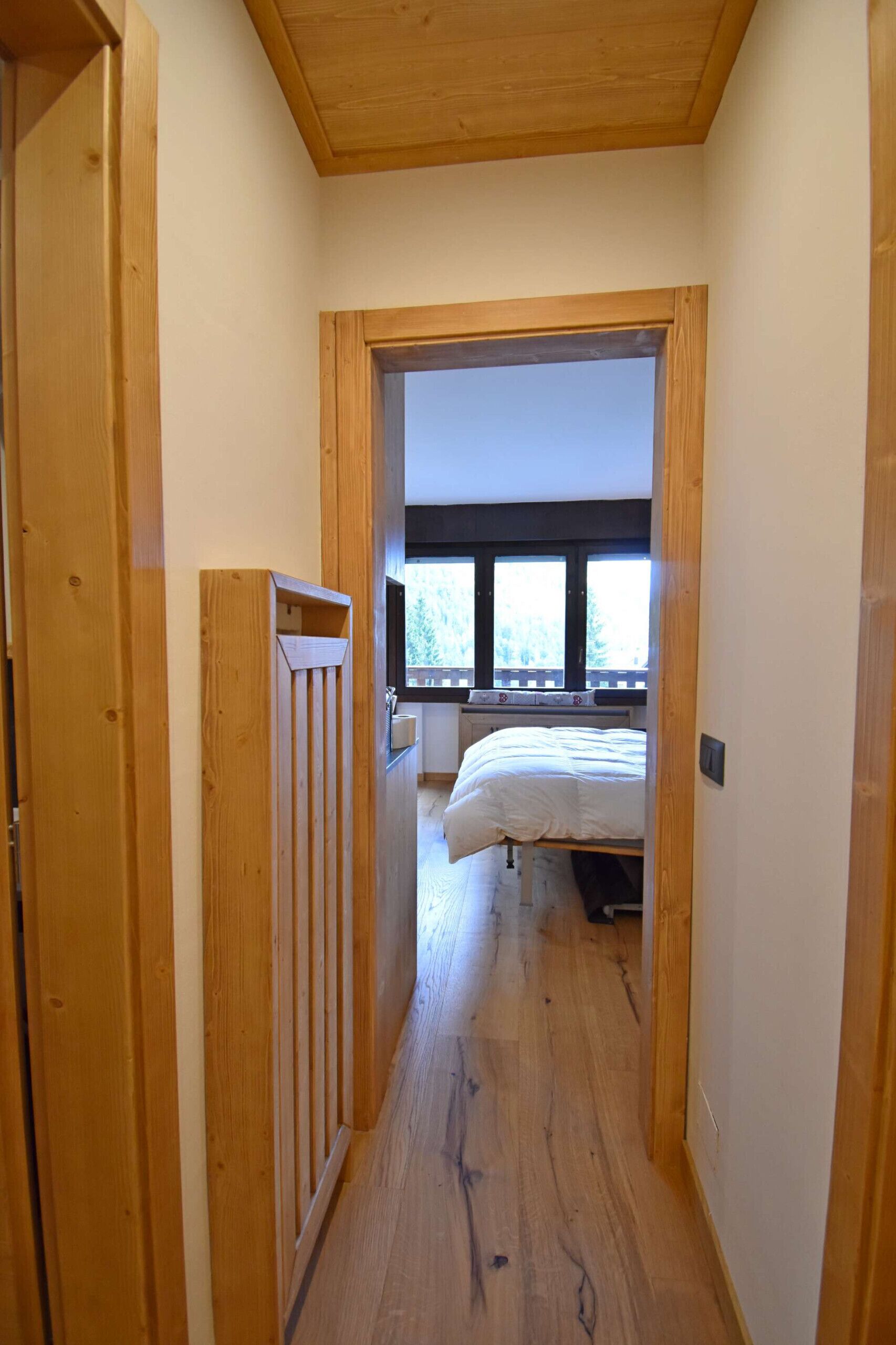 A narrow hallway with wooden walls and a wooden door on the left. A bed with white bedding is visible in the room at the end of the hallway.