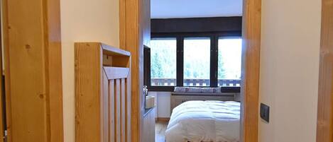 A narrow hallway with wooden walls and a wooden door on the left. A bed with white bedding is visible in the room at the end of the hallway.