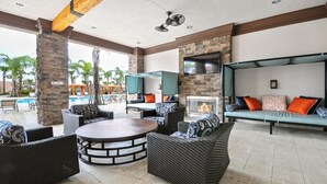 A spacious outdoor lounge area with wicker furniture, a round table, a fire pit, and a pool in the background.