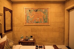 A hotel room with a large tapestry of an elephant on the wall, a wooden side table with toiletries, and a bed with a patterned throw. - Hotel Desert Castle Jaisalmer (Jaisalmer)