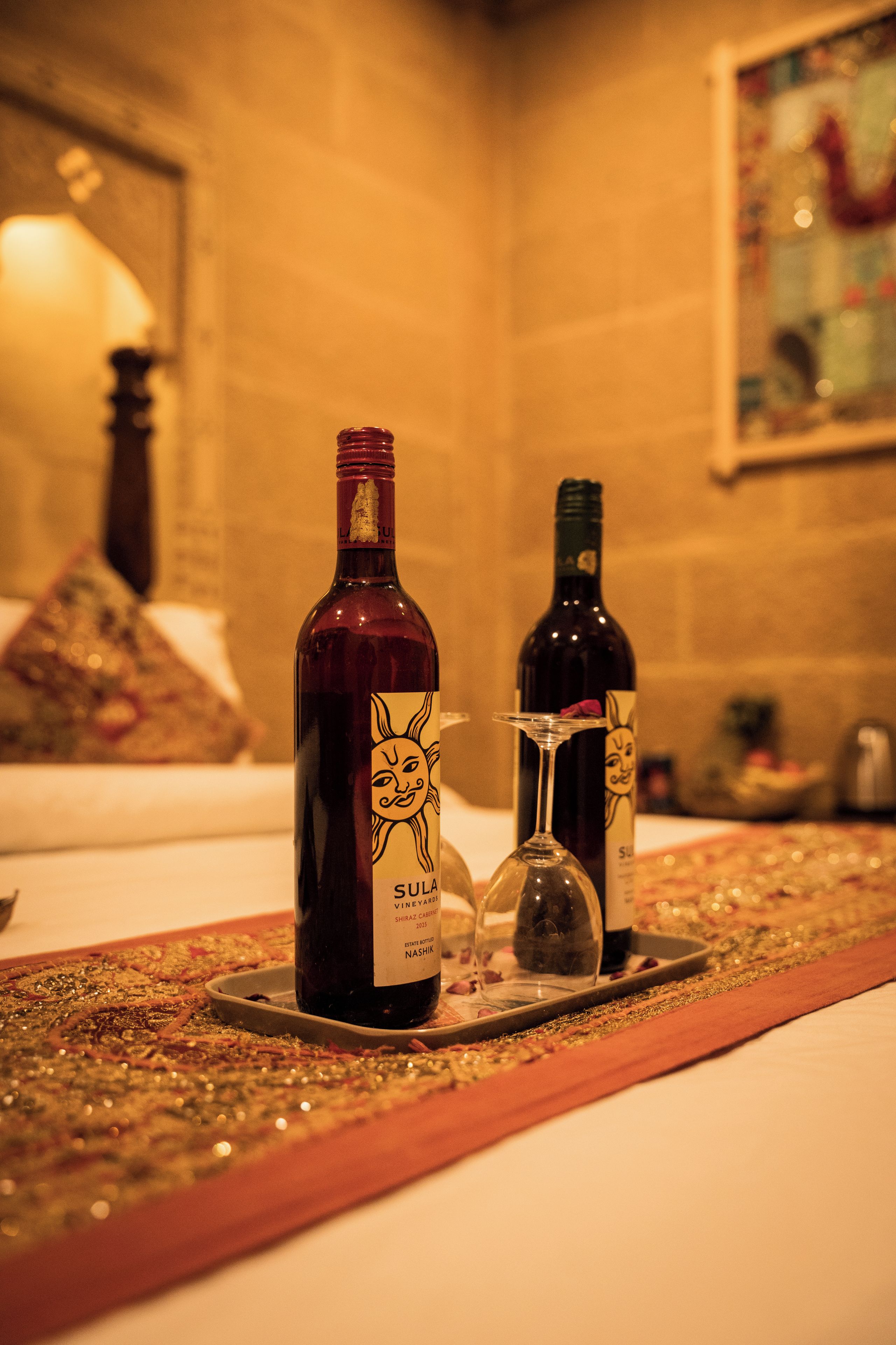 Uma garrafa de vinho tinto Shiraz Cabernet da Vinícola Sula, safra 2015, e uma taça de vinho em um tabuleiro com um jogo americano decorativo.