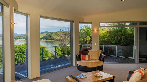 A spacious living area with a view of a lake and hills, featuring a sofa, a coffee table, and a chair. - Bay View Escape - Whitianga Holiday Home (Whitianga)