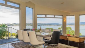 A dining area with a table set for four, large windows offering a view of a lake and mountains, and a balcony with a view of the lake. - Bay View Escape - Whitianga Holiday Home (Whitianga)