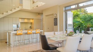 A modern kitchen with a central island, bar stools, and a dining area with a glass table and chairs. - Bay View Escape - Whitianga Holiday Home (Whitianga)