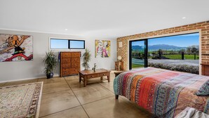 A bedroom with a large window, a bed with a colorful blanket, a dresser, and two wall-mounted TVs. - Kakepuku View and Spa Retreat - Te Awamutu (Te Awamutu)
