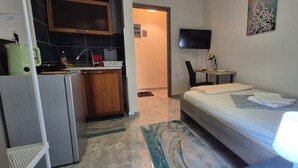 A compact hotel room with a bed, bedside lamp, a small table with a chair, a TV, and a kitchenette area with a fridge, microwave, and a kettle.