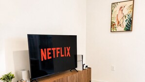 A modern living room with a large flat-screen TV, a wooden entertainment unit, a round ottoman, and a patterned rug. - Stylish Stay Near Hard Rock Stadium (North Miami Beach)