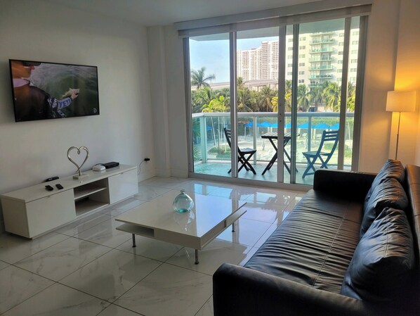 A modern living room with a flat-screen TV, a coffee table, a sofa, and a balcony with outdoor furniture. - Modern 1BR Condo on Sunny Isles across from the Beach and Free Parking on site (Sunny Isles Beach)