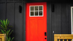 Una puerta roja con un ventanal, paredes exteriores negras y una planta pequeña a la izquierda.