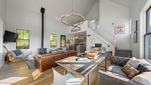 A spacious living area with a high ceiling, a fireplace, a dining table, and a staircase leading to an upper level.