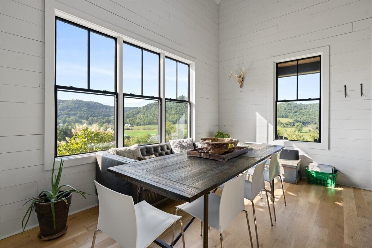 A dining area with a wooden table, white chairs, and a bench. Large windows offer a view of a green landscape.