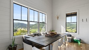 A dining area with a wooden table, white chairs, and a bench. Large windows offer a view of a green landscape.