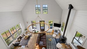 A spacious loft-style living area with a high ceiling, large windows, a central chandelier, and a built-in fireplace.