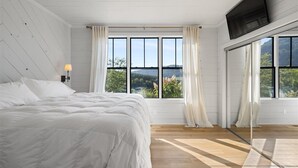A bedroom with a large bed, a TV mounted on the wall, and a view of the outdoors through large windows.