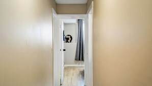Un couloir étroit aux murs de couleur beige et au plancher en bois, qui mène à une pièce dotée d’un rideau de douche et d’un élément décoratif mural.